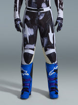 Alpinestars 2026 Racer Lahnd Pants, pantaloni da motocross, Iron Camo, indossati con guanti mimetici coordinati e stivali blu acceso e viola, con grafiche a stella bianche stilizzate su motivo mimetico scuro, chiusura con fibbia magnetica tecnica, abbigliamento professionale da fuoristrada