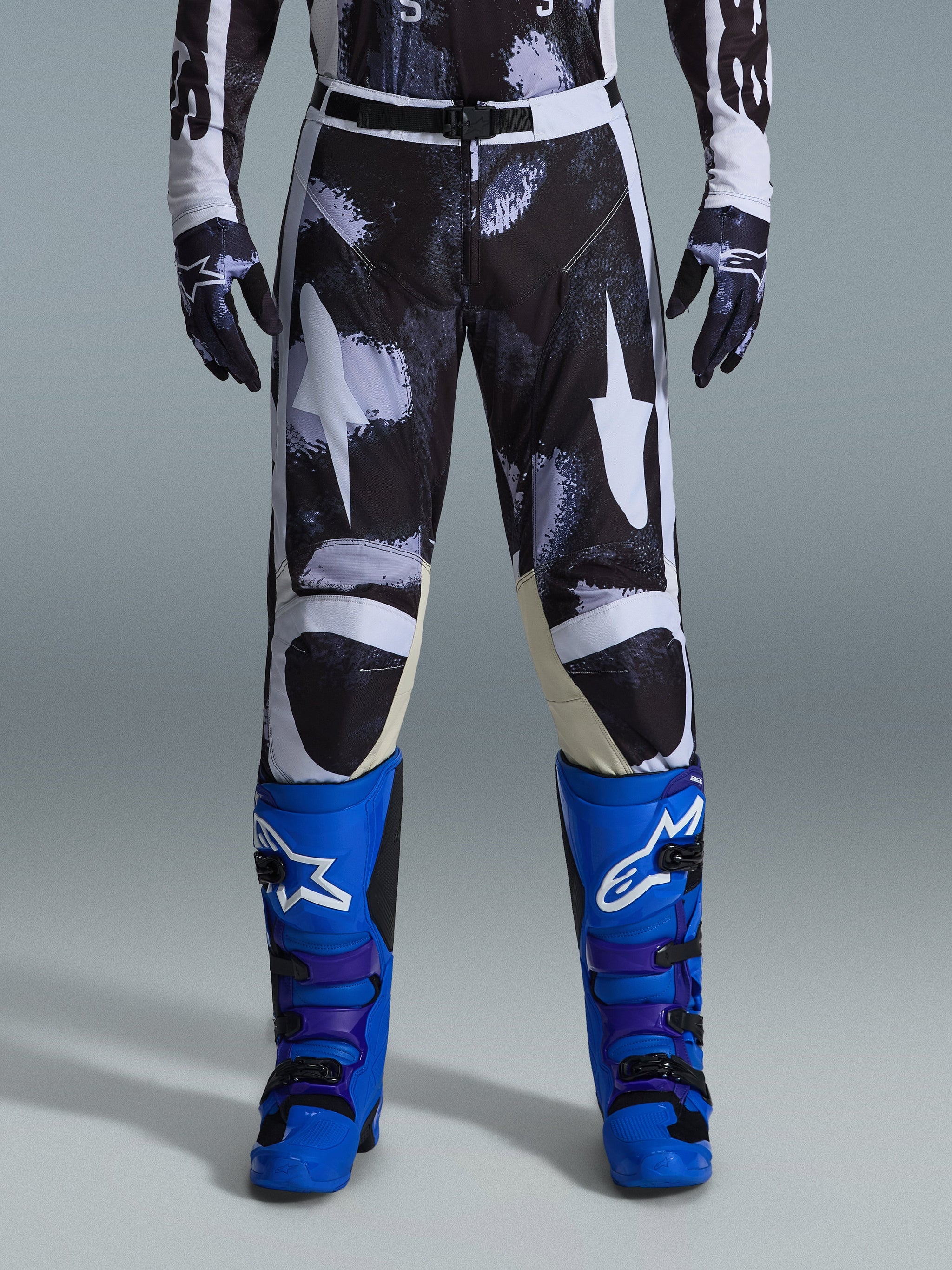 Alpinestars 2026 Racer Lahnd Pants, pantaloni da motocross, Iron Camo, indossati con guanti mimetici coordinati e stivali blu acceso e viola, con grafiche a stella bianche stilizzate su motivo mimetico scuro, chiusura con fibbia magnetica tecnica, abbigliamento professionale da fuoristrada