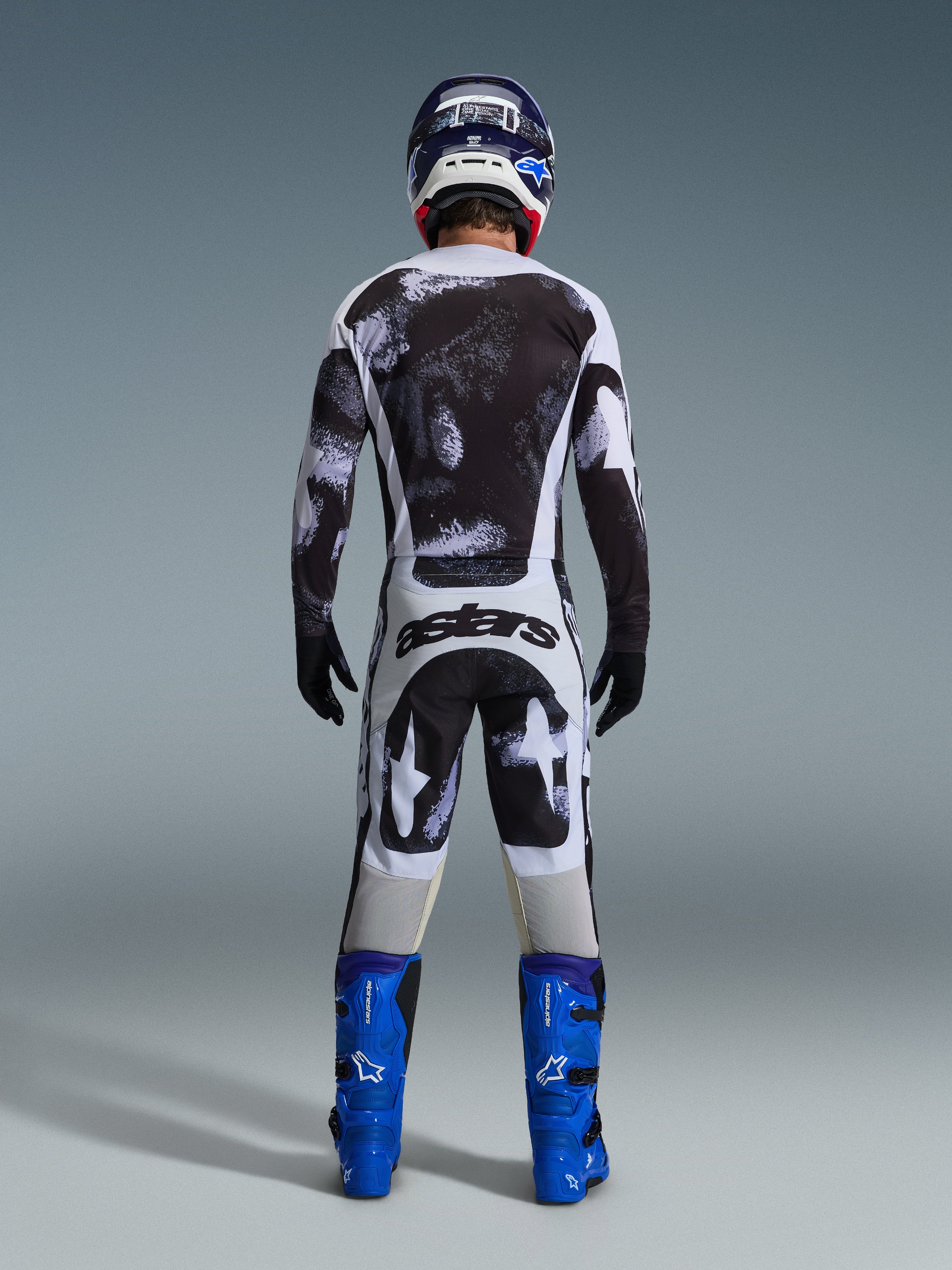 Pilota di motocross visto da dietro che indossa la maglia Alpinestars Racer Lahnd 2026 e i pantaloni coordinati in Iron Camo, caratterizzati da un motivo splatter bianco e nero, abbinati a stivali blu acceso e casco coordinato, set completo per corse fuoristrada