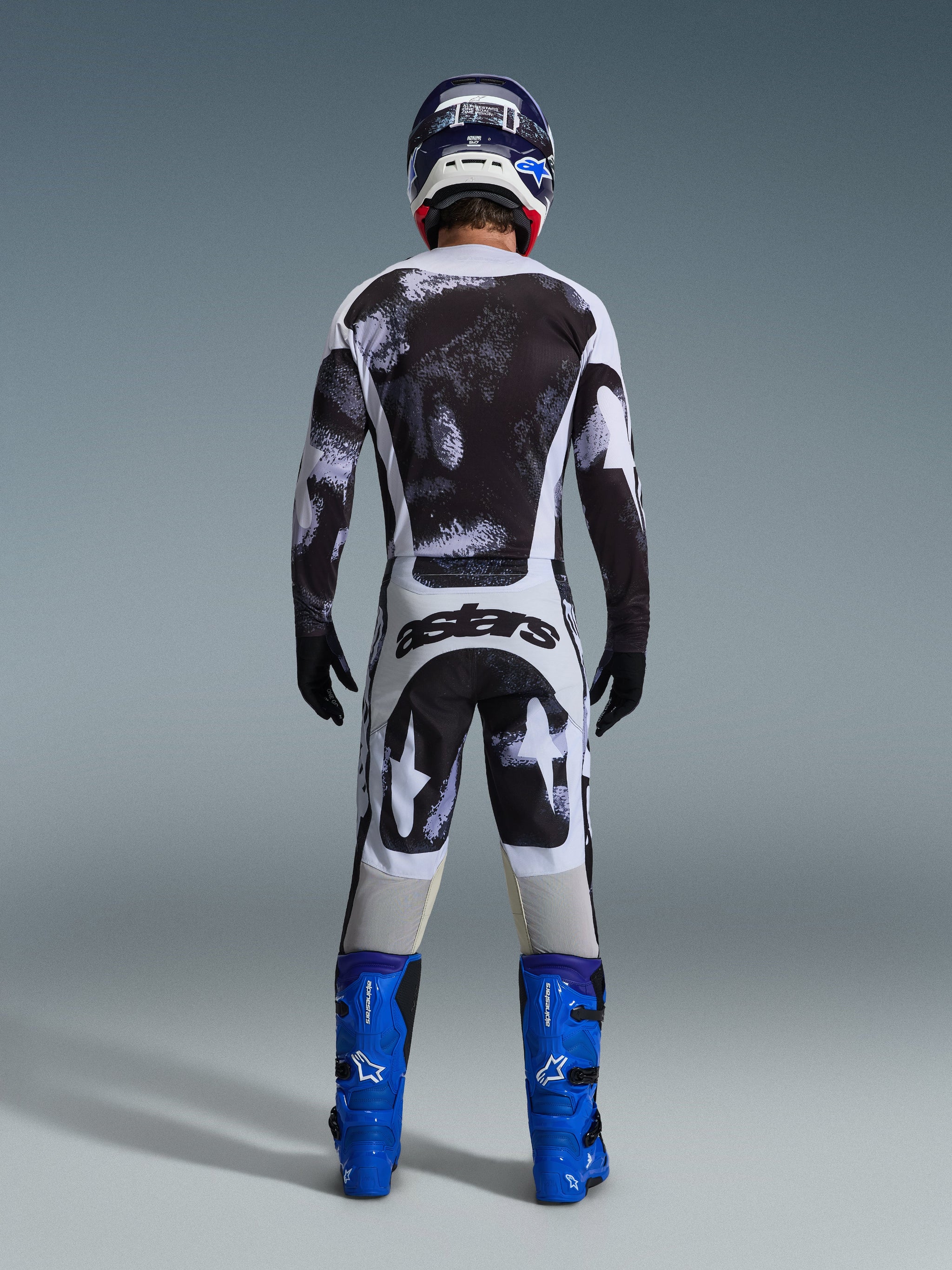 Alpinestars 2026 Racer Lahnd Pants, pantaloni da motocross, motivo Iron Camo grigio e nero, vista posteriore del pilota in tenuta da gara completa con maglia coordinata e stivali blu, progettati per prestazioni off-road