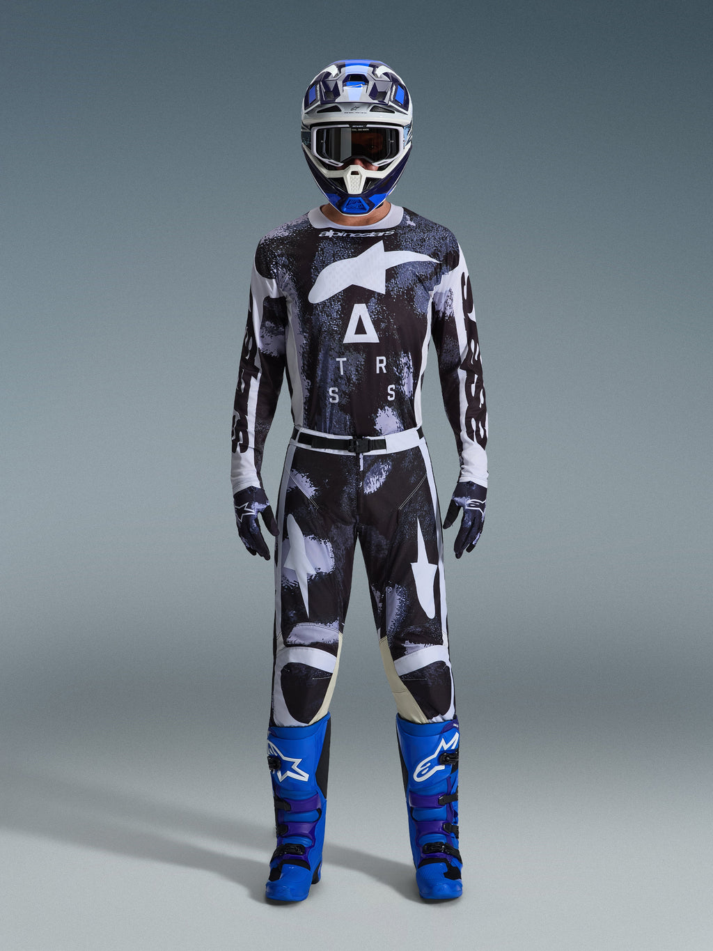 Pilota di motocross con pantaloni Alpinestars Racer Lahnd 2026 in Iron Camo, design mimetico grigio e nero, maglia coordinata, casco blu e bianco con occhiali e stivali blu acceso, set di abbigliamento da motocross coordinato.