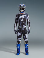 Pilota di motocross con pantaloni Alpinestars Racer Lahnd 2026 in Iron Camo, design mimetico grigio e nero, maglia coordinata, casco blu e bianco con occhiali e stivali blu acceso, set di abbigliamento da motocross coordinato.