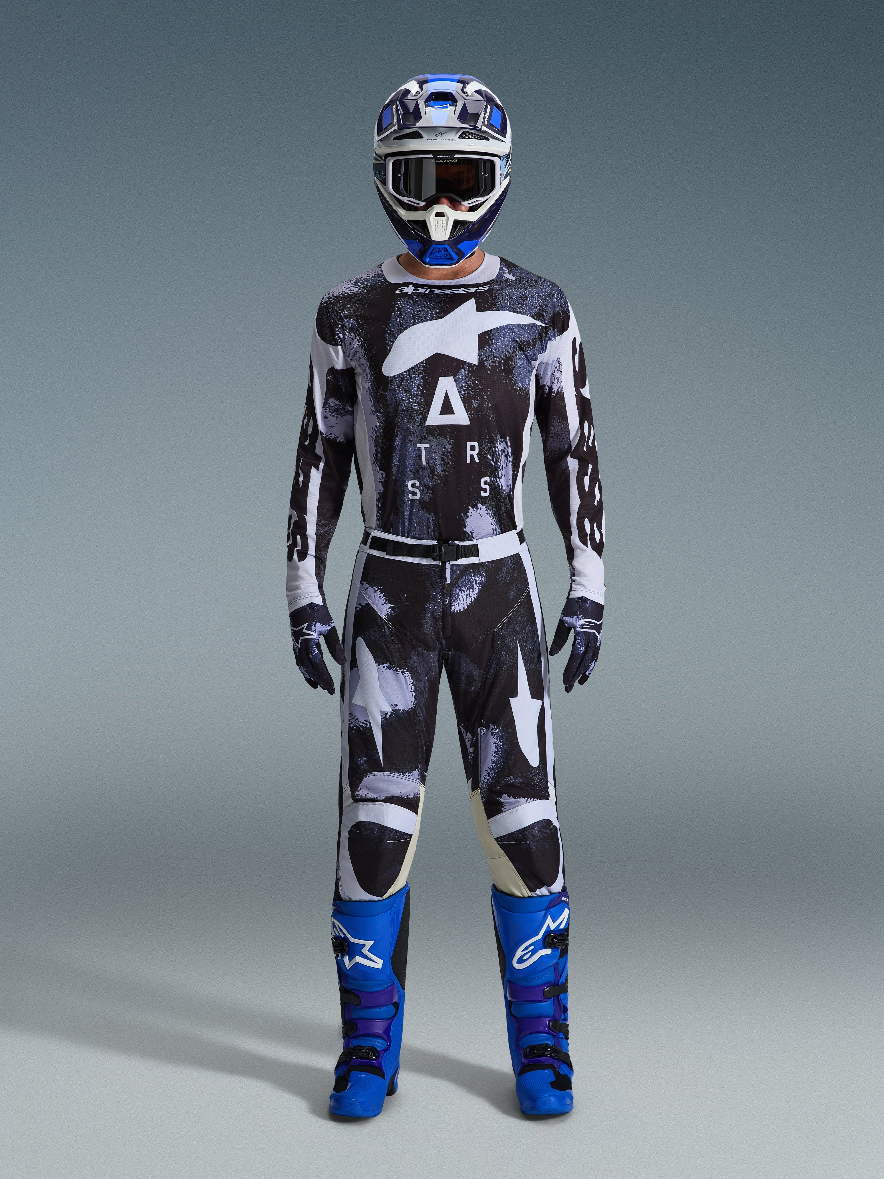 Pilota di motocross con pantaloni Alpinestars Racer Lahnd 2026 in Iron Camo, design mimetico grigio e nero, maglia coordinata, casco blu e bianco con occhiali e stivali blu acceso, set di abbigliamento da motocross coordinato.