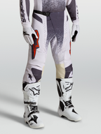 Alpinestars Pantaloni Techstar Swan, pantaloni da motocross da uomo, lilla chiaro e grigio scuro con dettagli rosso fluo, indossati con guanti e stivali bianchi, abbigliamento tecnico per off-road e competizioni