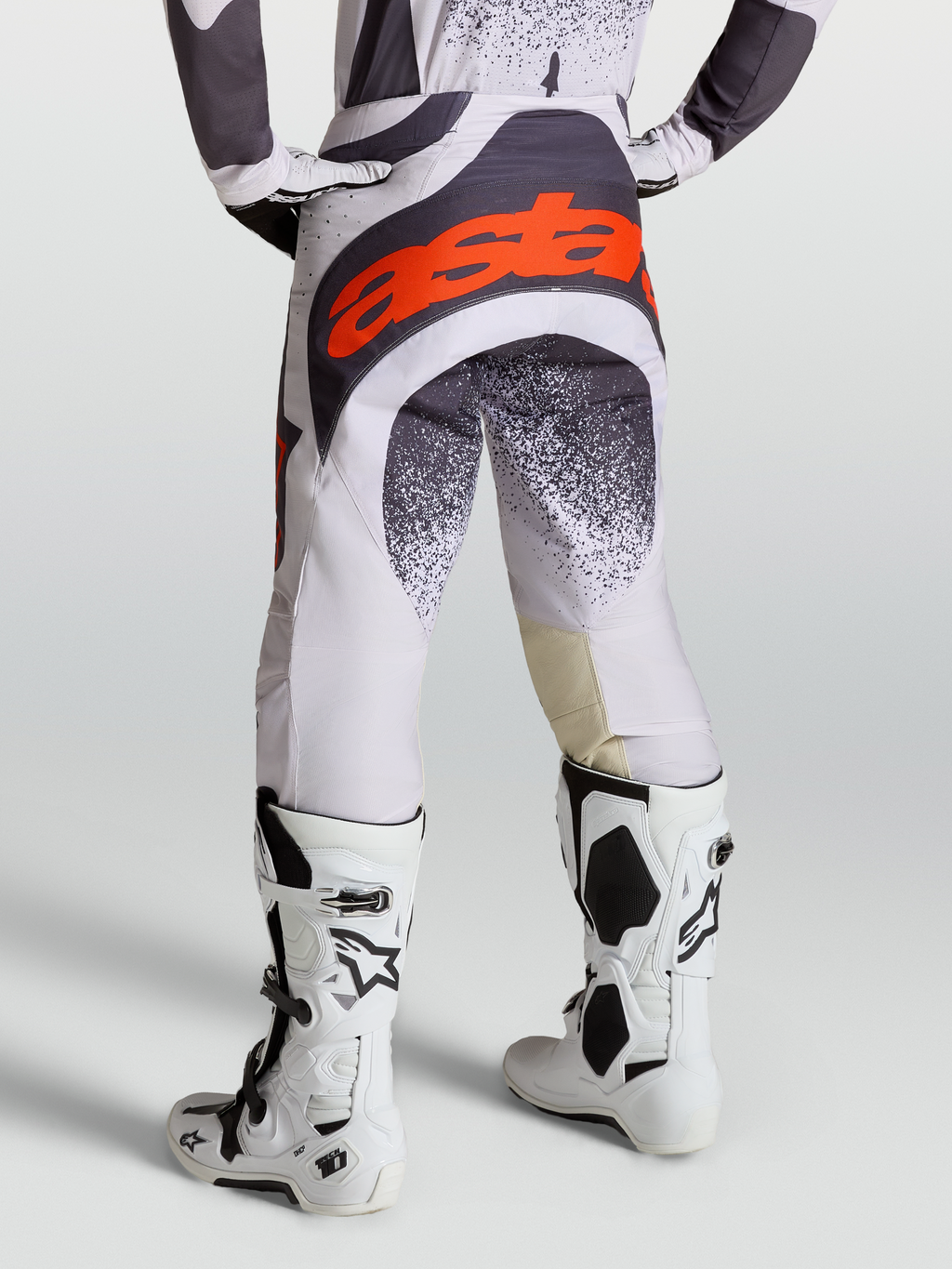 Alpinestars Pantaloni Techstar Swan, pantaloni da motocross grigi e lilla per uomo, con loghi rosso fluo e abbinati a stivali Tech 10 bianchi, vista posteriore su modello.