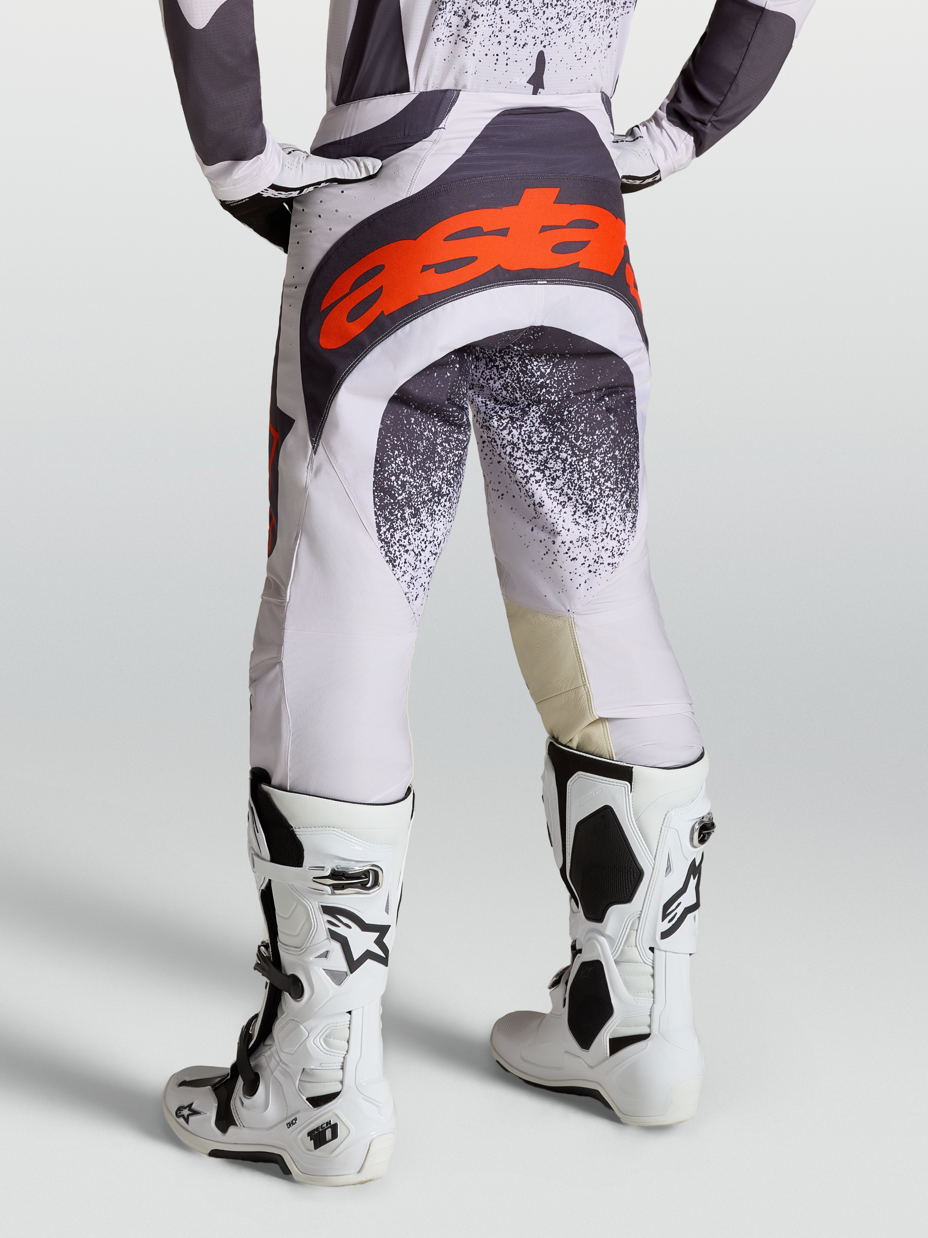 Alpinestars Pantaloni Techstar Swan, pantaloni da motocross grigi e lilla per uomo, con loghi rosso fluo e abbinati a stivali Tech 10 bianchi, vista posteriore su modello.