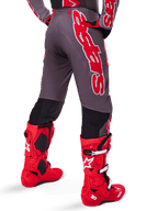 Alpinestars 2025 Supertech Lipan Gear Set, vista posteriore di un pilota di motocross, pantaloni grigio scuro con grande logo verticale rosso, maglia coordinata rossa e grigia, stivali da motocross rosso acceso, abbigliamento da gara fuoristrada ad alte prestazioni