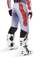 Pantaloni Alpinestars Supertech Lipan 2025, pantaloni da motocross, grigio e arancione fluo, indossati con maglia coordinata e stivali bianchi, vista posteriore con grande grafica arancione del marchio Alpinestars, pannelli elasticizzati neri, abbigliamento professionale per motocross