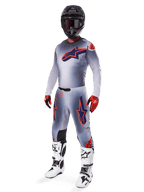 Pilota di motocross con completo da gara Alpinestars Supertech Lipan 2025, grigio e arancione fluo, maglia grigia con grande logo arancione sul petto, pantaloni coordinati con accenti arancioni, casco nero, guanti rossi e stivali bianchi, abbigliamento professionale da gara fuoristrada