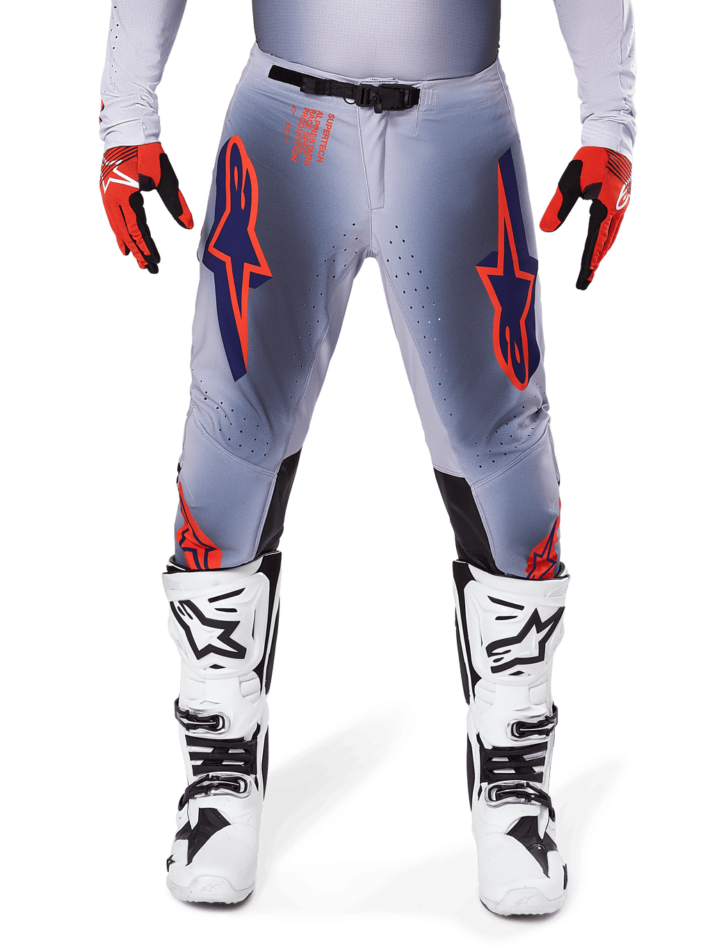 Pantaloni Alpinestars Supertech Lipan 2025, pantaloni da motocross, grigio e arancione fluo, indossati con guanti rossi e neri e stivali bianchi e neri, con grandi loghi a stella Alpinestars sulle cosce e perforazioni per la ventilazione per gare di livello professionale.