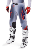 Pantaloni Alpinestars Supertech Lipan 2025, pantaloni da motocross, grigio e arancione fluo, con design a stelle blu navy e pannelli interni neri sulle ginocchia, indossati con guanti rosso-arancio e stivali bianchi, abbigliamento professionale da gara fuoristrada