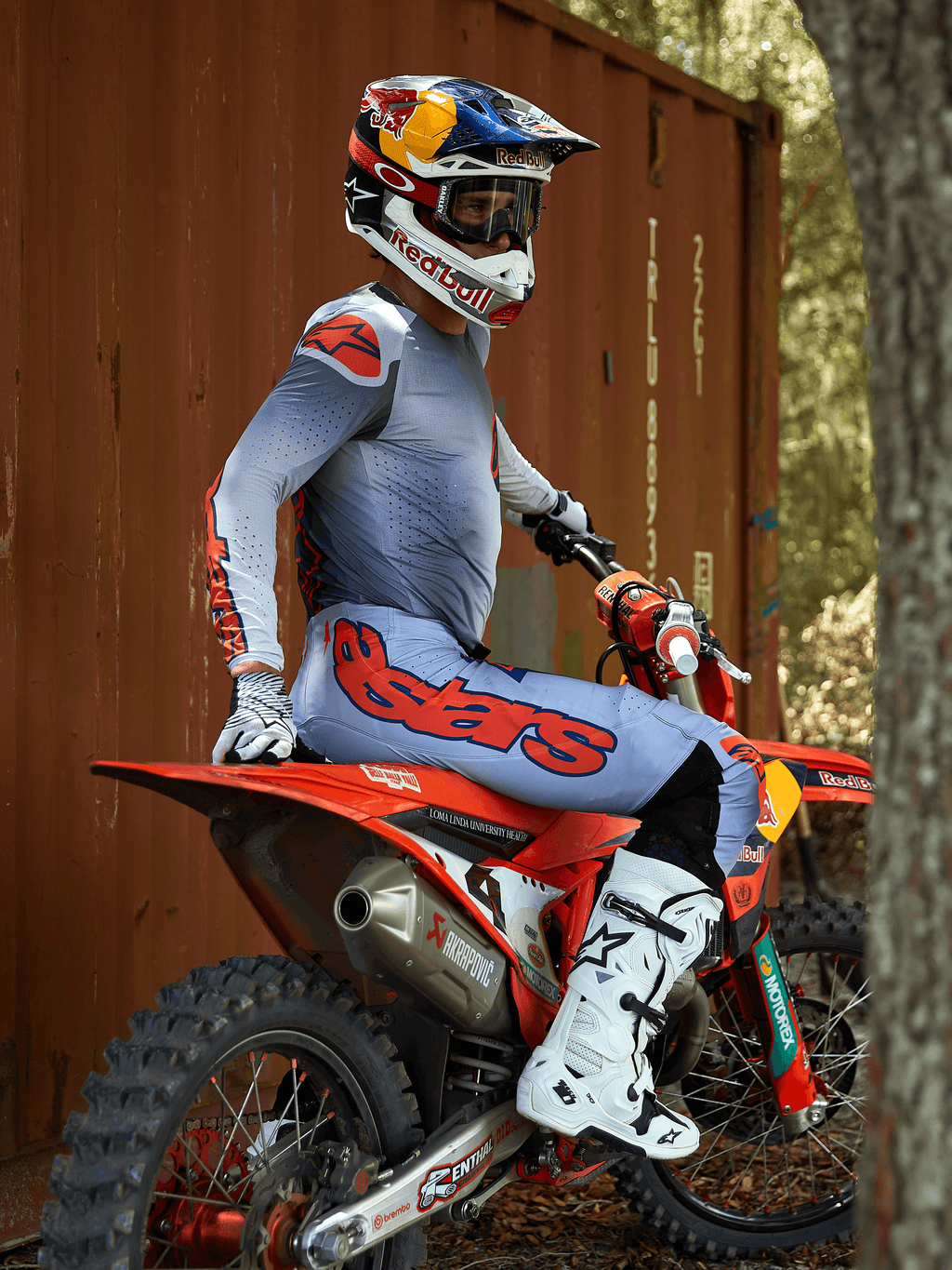 Pilota di motocross seduto su una moto da cross rossa, indossa il completo Alpinestars 2025 Supertech Lipan in Grigio e Arancio Fluo, pantaloni azzurro chiaro con vistoso logo Astars arancione, maglia coordinata, stivali da motocross bianchi e casco Red Bull, abbigliamento professionale da gara