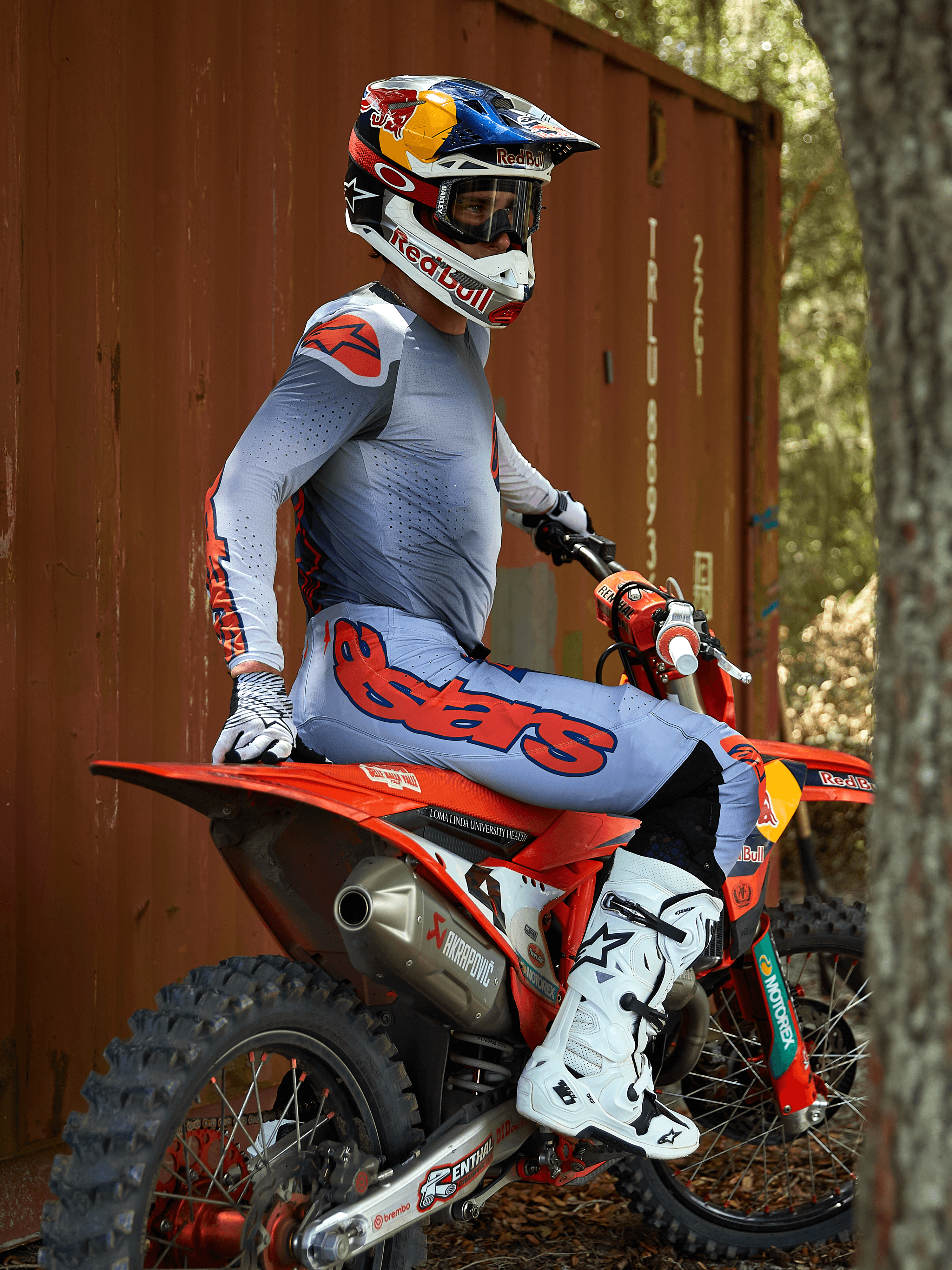 Pilota di motocross seduto su una moto da cross rossa, indossa il completo Alpinestars 2025 Supertech Lipan in Grigio e Arancio Fluo, pantaloni azzurro chiaro con vistoso logo Astars arancione, maglia coordinata, stivali da motocross bianchi e casco Red Bull, abbigliamento professionale da gara