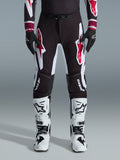 Alpinestars 2026 Supertech AFD Vista Pants, pantaloni da motocross, nero rosso e bianco, con grandi loghi grafici sulle cosce e sulle ginocchia, indossati con stivali da gara bianchi e guanti neri, design atletico race-ready per competizioni off-road d'élite