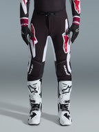 Alpinestars 2026 Supertech AFD Vista Pants, pantaloni da motocross, nero rosso e bianco, con grandi loghi grafici sulle cosce e sulle ginocchia, indossati con stivali da gara bianchi e guanti neri, design atletico race-ready per competizioni off-road d'élite
