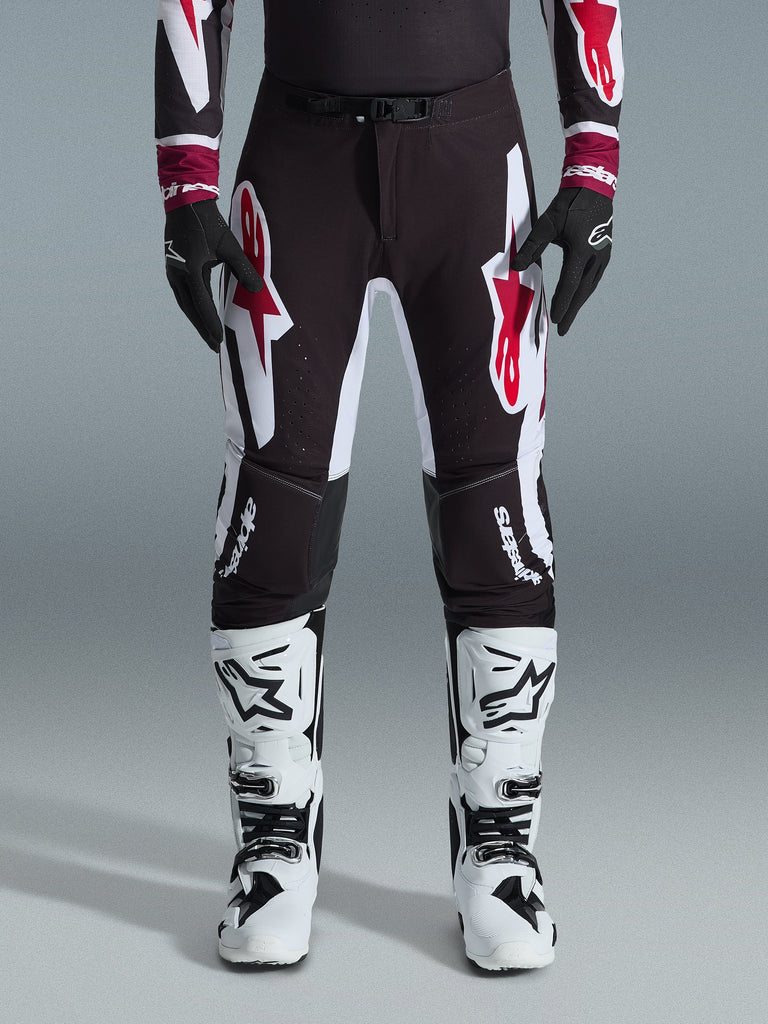 Alpinestars 2026 Supertech AFD Vista Pants, pantaloni da motocross, nero rosso e bianco, con grandi loghi grafici sulle cosce e sulle ginocchia, indossati con stivali da gara bianchi e guanti neri, design atletico race-ready per competizioni off-road d'élite