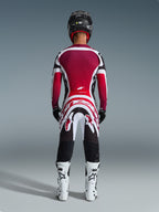 Pilota di motocross visto da dietro che indossa pantaloni Alpinestars 2026 Supertech AFD Vista, Nero Rosso Bianco, abbinati a una maglia coordinata, stivali da gara bianchi e casco nero, set completo di abbigliamento professionale per gare fuoristrada
