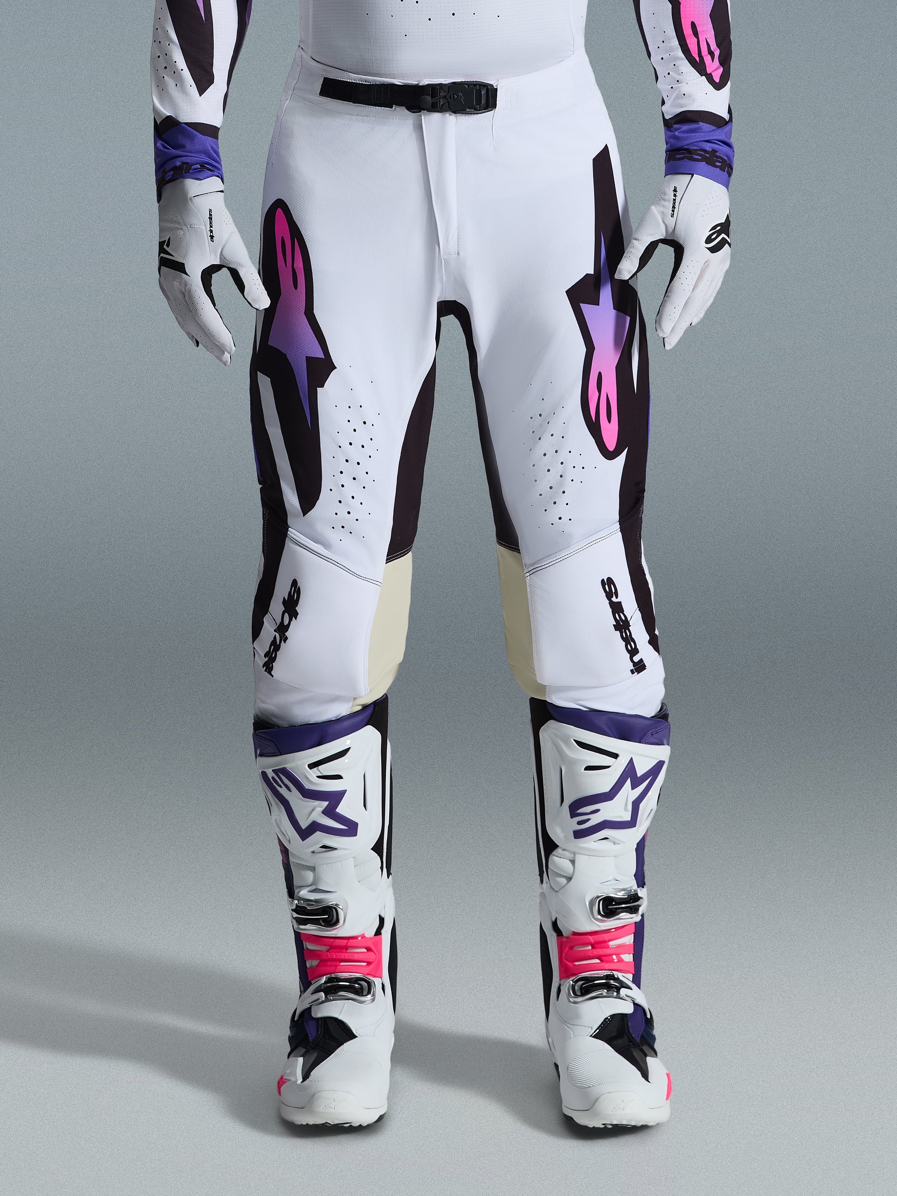 Alpinestars 2026 Supertech AFD Vista Gear Set, pilota di motocross con completo da gara bianco e accenti viola e rosa, caratterizzato da pantaloni ad alte prestazioni con vistosi loghi a stella, maglia e guanti coordinati e stivali da motocross bianchi con dettagli rosa e viola.