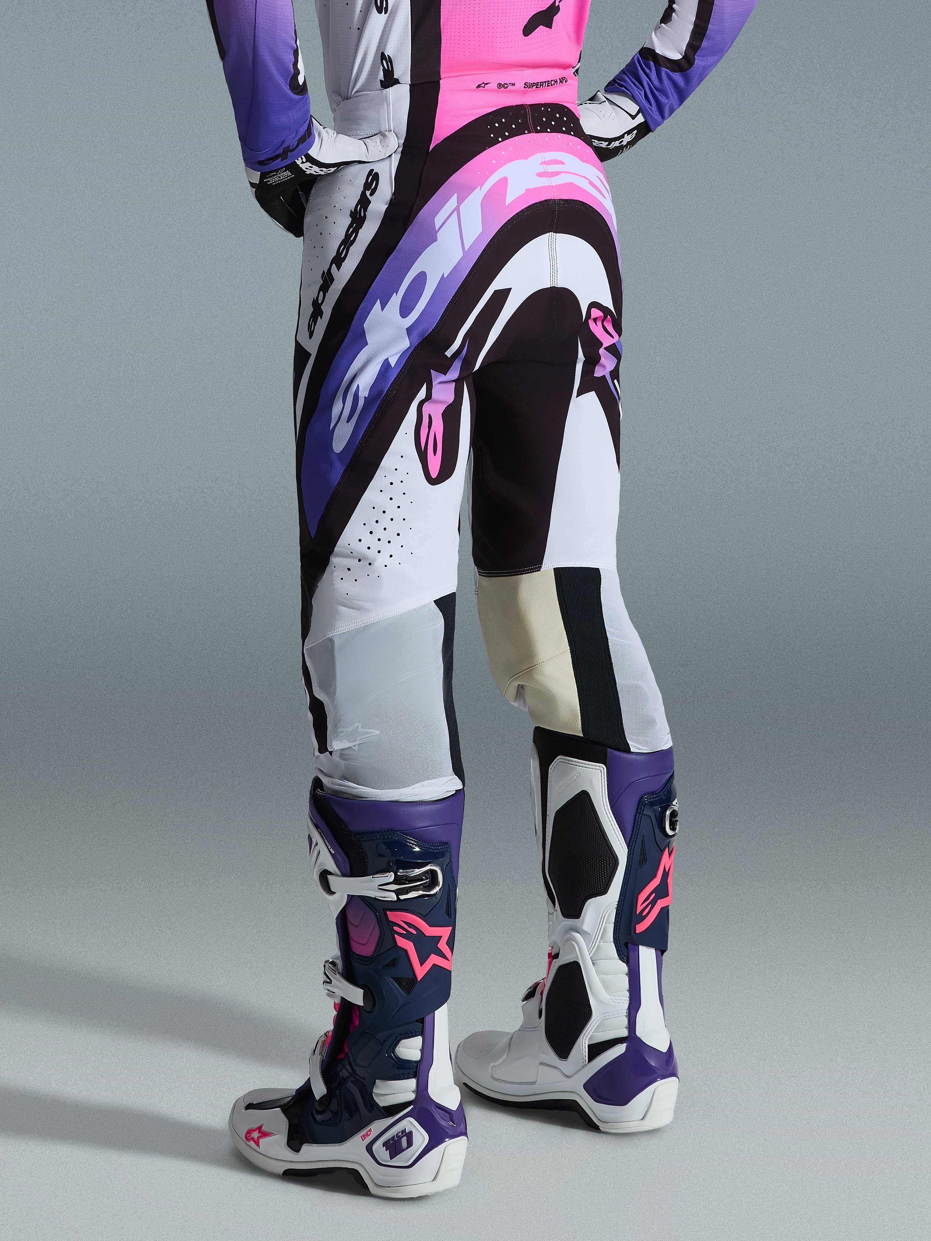 Pantaloni Alpinestars 2026 Supertech AFD Vista, pantaloni da motocross, bianco, viola e nero con accenti rosa, vista posteriore, indossati con maglia coordinata e stivali Tech 10, con grafiche logo audaci e materiali performanti per gare off-road d'élite