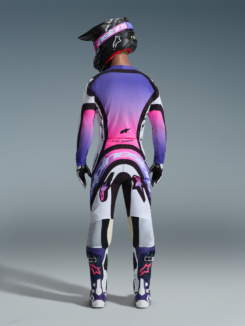 Pilota di motocross visto da dietro con abbigliamento da gara completo, maglia con sfumatura viola e rosa, pantaloni Alpinestars 2026 Supertech AFD Vista in bianco, viola e nero, casco nero con maschera viola, stivali bianchi e viola, kit da gara off-road coordinato.