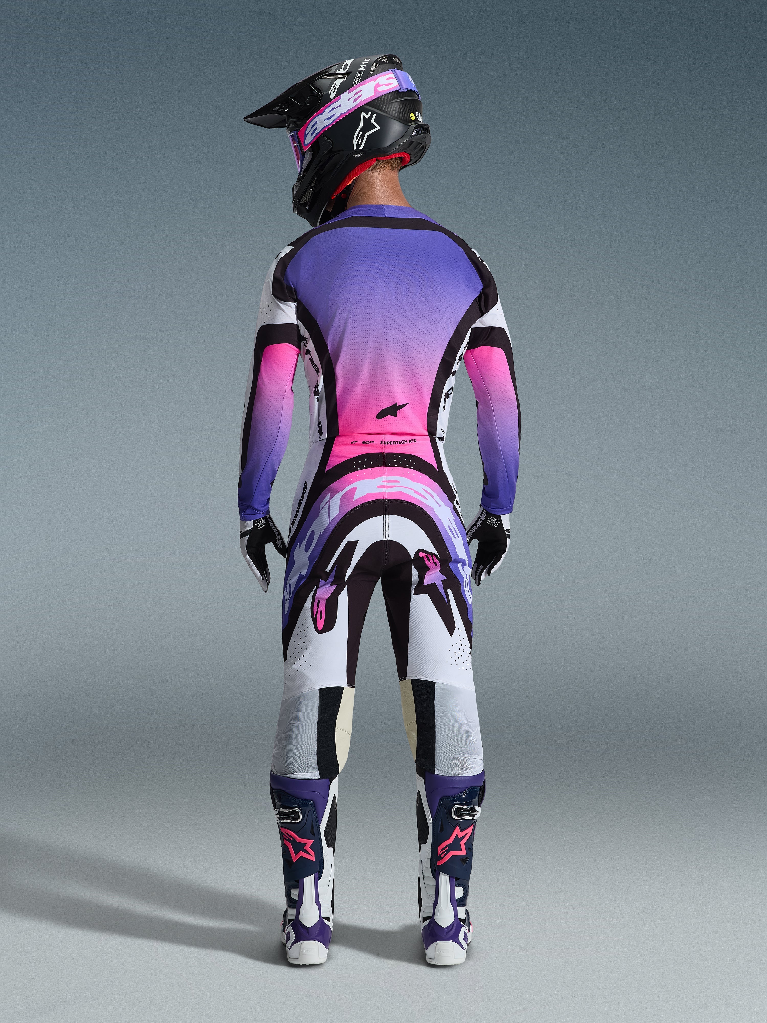 Pilota di motocross visto da dietro con abbigliamento da gara completo, maglia con sfumatura viola e rosa, pantaloni Alpinestars 2026 Supertech AFD Vista in bianco, viola e nero, casco nero con maschera viola, stivali bianchi e viola, kit da gara off-road coordinato.