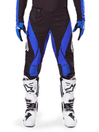 Alpinestars 2025 Techstar Dreem Pants, pantaloni da motocross, nero e blu, indossati con maglia e guanti coordinati e stivali bianchi, con pannelli laterali blu e vestibilità atletica da gara, materiale resistente per corse fuoristrada