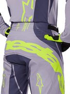 Alpinestars 2025 Techstar Dreem Pants, pantaloni da motocross, grigio e grigio scuro con accenti giallo fluorescente, vista posteriore con marchio Alpinestars sul carré, indossati con maglia e guanti coordinati, progettati per gare fuoristrada