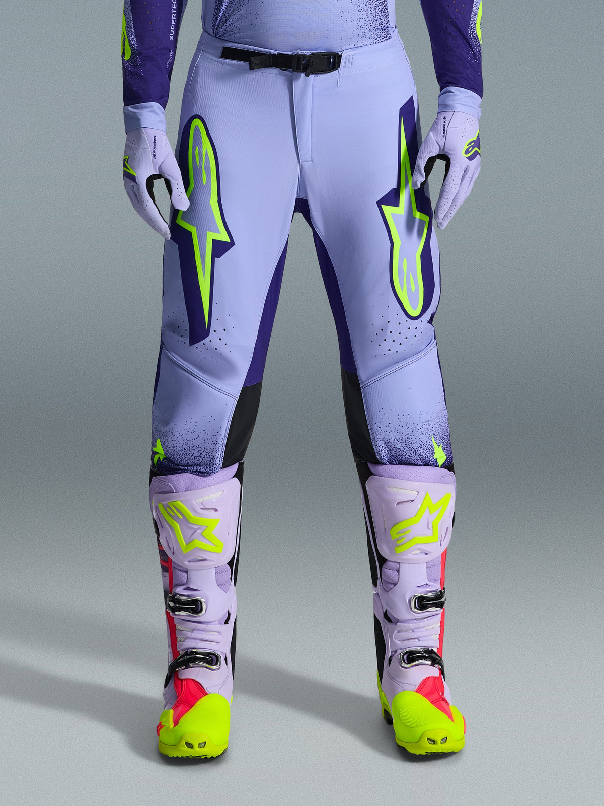 Alpinestars 2026 Supertech Scenz Pants, pantaloni da motocross, viola giallo fluo, con grandi loghi giallo neon sulle cosce, indossati con stivali e guanti da motocross coordinati viola e gialli, design performante con vestibilità atletica per corse fuoristrada