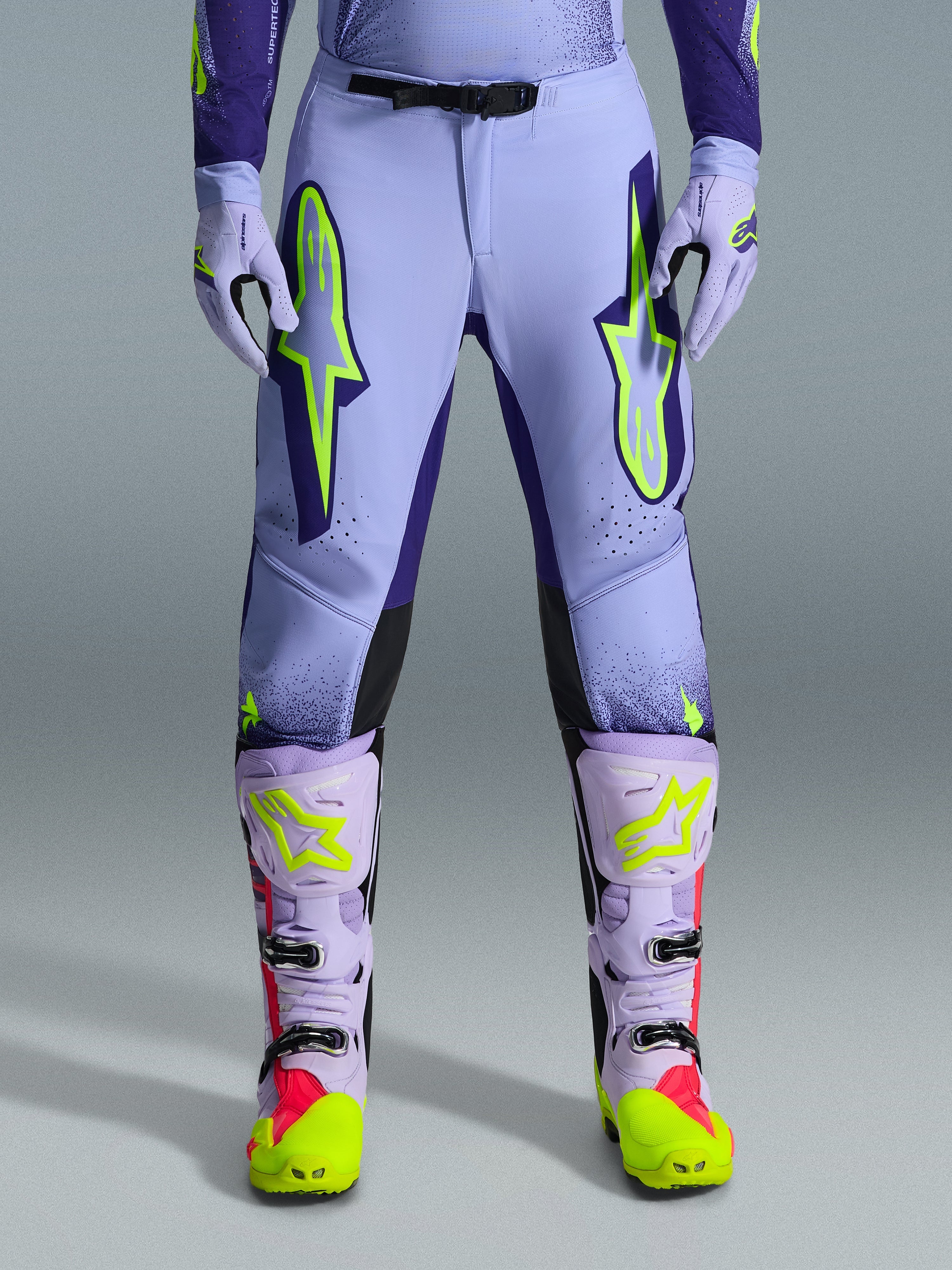 Alpinestars 2026 Supertech Scenz Pants, pantaloni da motocross, viola giallo fluo, con grandi loghi giallo neon sulle cosce, indossati con stivali e guanti da motocross coordinati viola e gialli, design performante con vestibilità atletica per corse fuoristrada