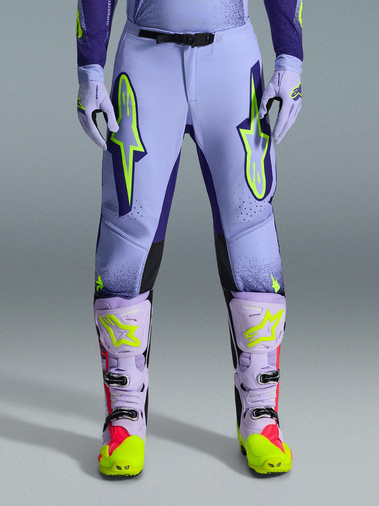 Alpinestars 2026 Supertech Scenz Pants, pantaloni da motocross, viola giallo fluo, con grandi loghi giallo neon sulle cosce, indossati con stivali e guanti da motocross coordinati viola e gialli, design performante con vestibilità atletica per corse fuoristrada