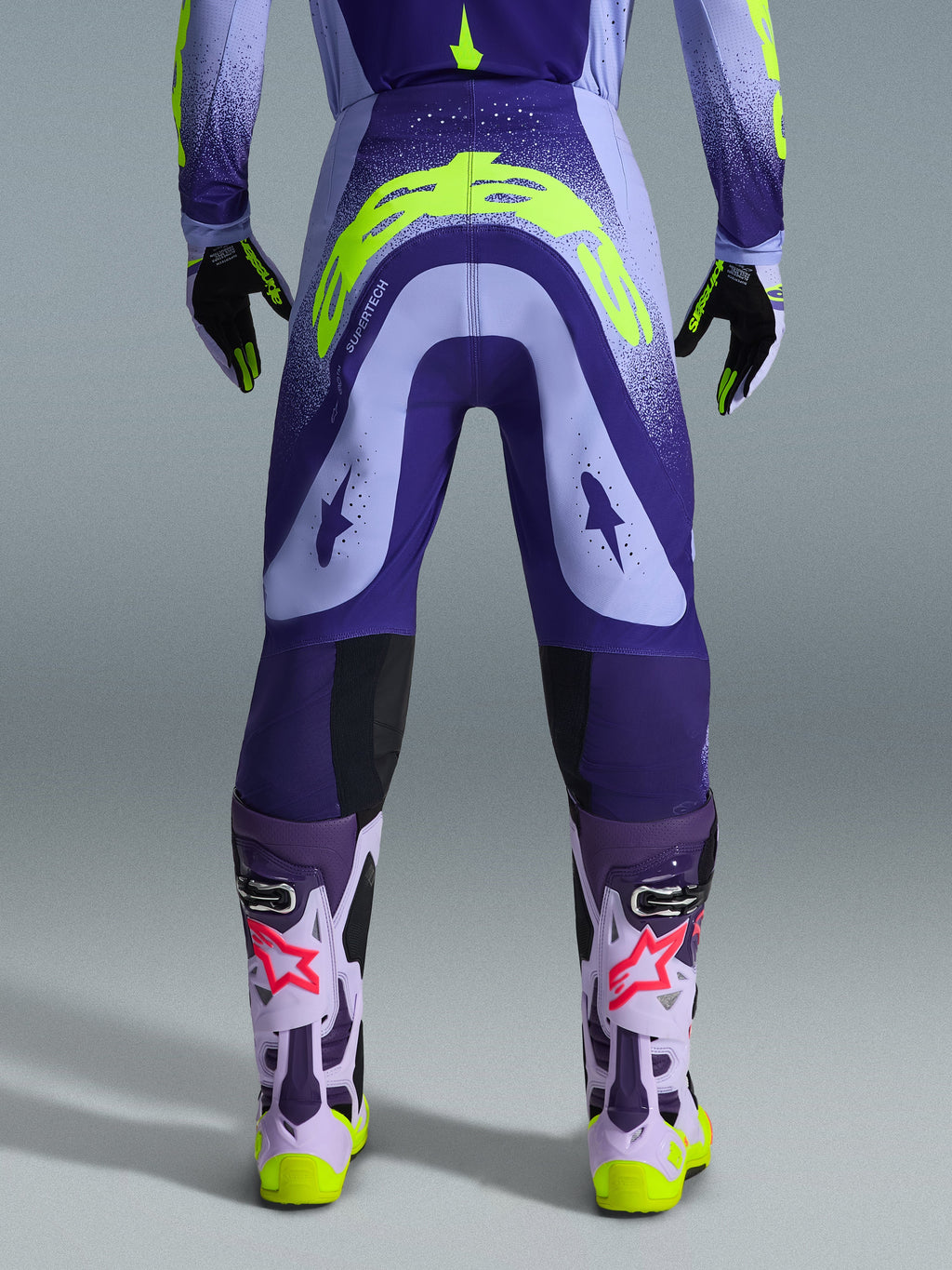 Alpinestars 2026 Supertech Scenz Pants, pantaloni da motocross, viola e giallo fluo, vista posteriore con grande logo giallo sulla seduta, indossati con maglia, guanti e stivali protettivi coordinati, vestibilità atletica per gare di motocross professionistiche