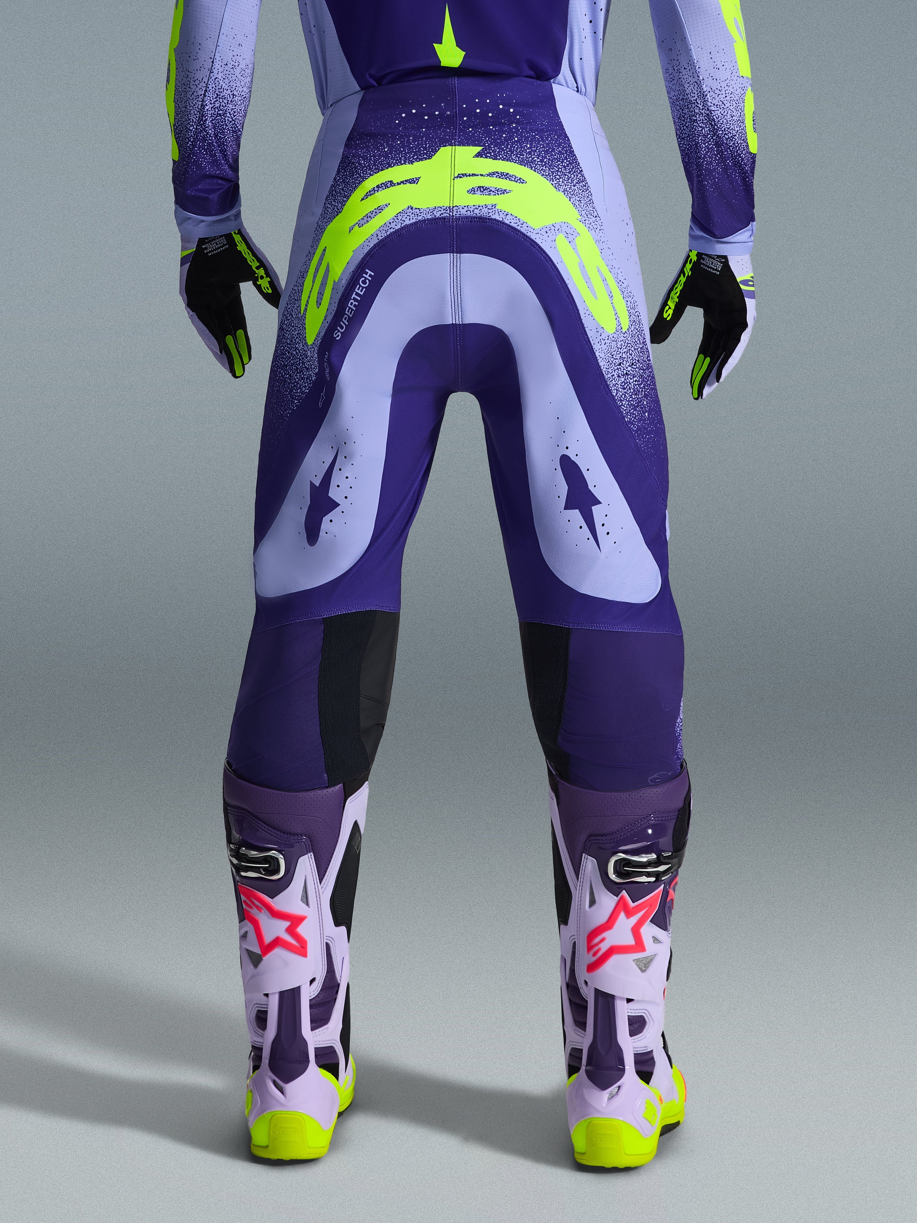 Alpinestars 2026 Supertech Scenz Pants, pantaloni da motocross, viola e giallo fluo, vista posteriore con grande logo giallo sulla seduta, indossati con maglia, guanti e stivali protettivi coordinati, vestibilità atletica per gare di motocross professionistiche