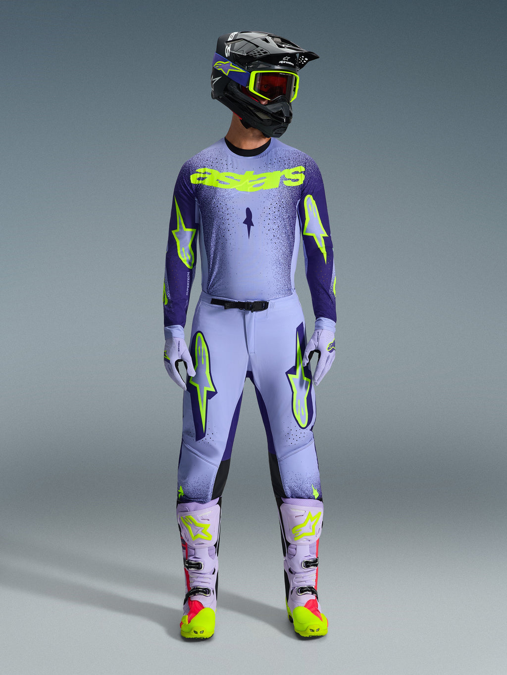 Alpinestars 2026 Supertech Scenz Gear Set, completo motocross, viola chiaro e giallo-verde neon, equipaggiamento protettivo completo comprensivo di casco nero con accenti viola, maglia con branding giallo neon, pantaloni coordinati con grafiche a stella e stivali coordinati con dettagli rosa.