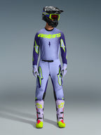 Alpinestars 2026 Supertech Scenz Gear Set, completo motocross, viola chiaro e giallo-verde neon, equipaggiamento protettivo completo comprensivo di casco nero con accenti viola, maglia con branding giallo neon, pantaloni coordinati con grafiche a stella e stivali coordinati con dettagli rosa.