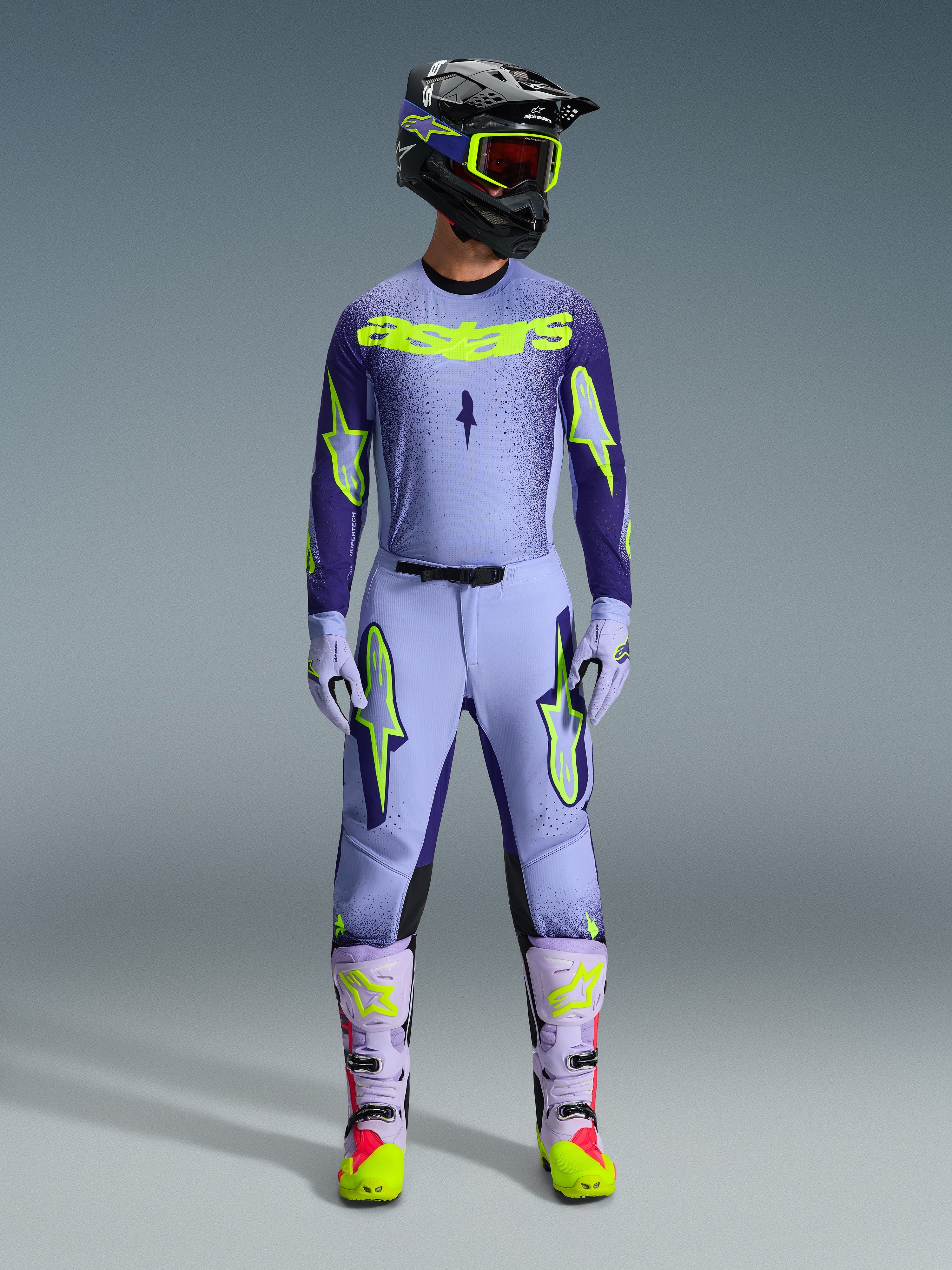 Alpinestars 2026 Supertech Scenz Gear Set, completo motocross, viola chiaro e giallo-verde neon, equipaggiamento protettivo completo comprensivo di casco nero con accenti viola, maglia con branding giallo neon, pantaloni coordinati con grafiche a stella e stivali coordinati con dettagli rosa.