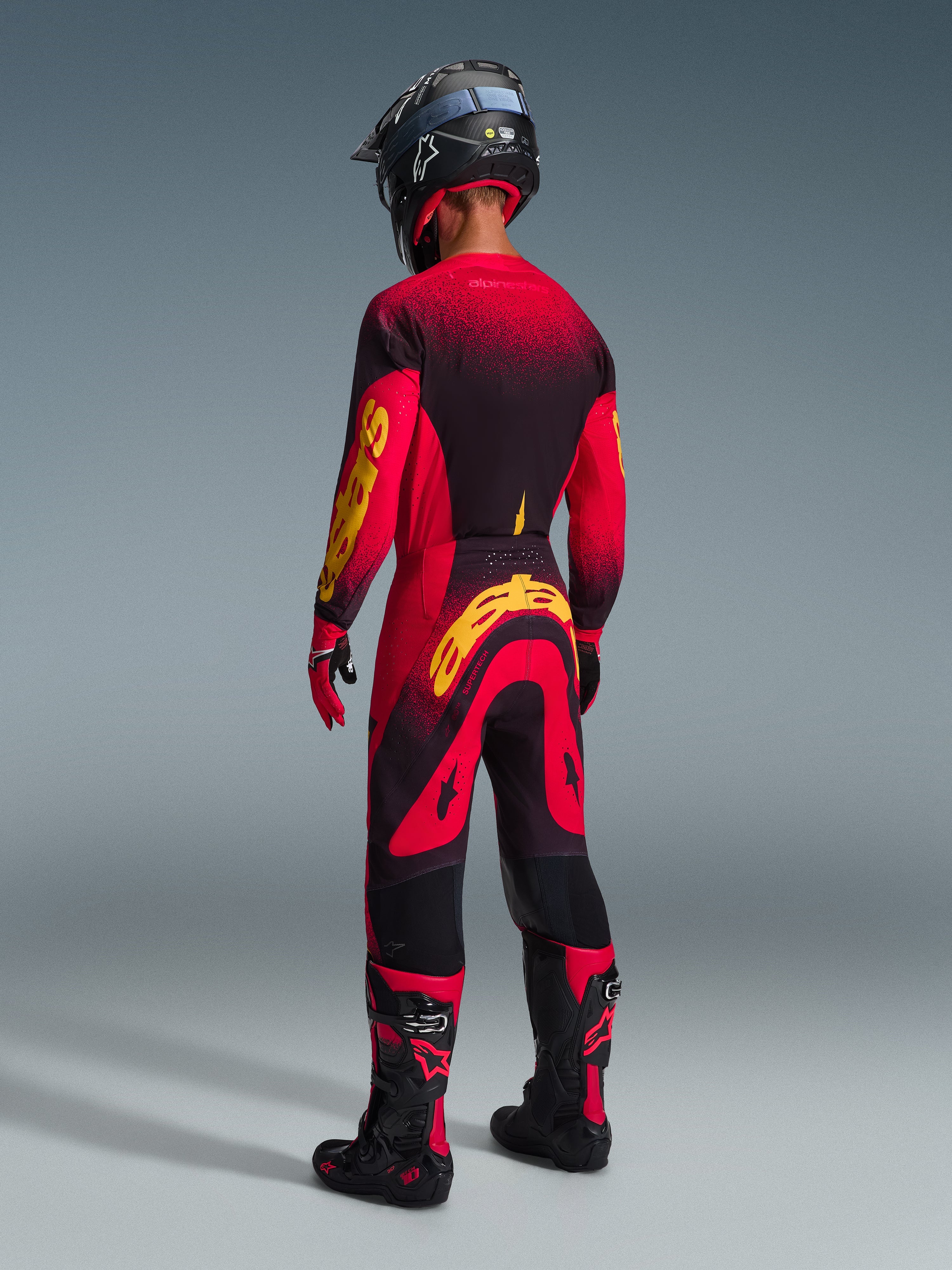 Pilota di motocross visto da dietro che indossa pantaloni Alpinestars 2026 Supertech Scenz in Rosso Nero Giallo, maglia coordinata con logo giallo, stivali da motocross neri e rossi, casco nero, equipaggiamento professionale completo per performance fuoristrada.