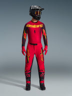 Alpinestars 2026 Supertech Scenz Pants, pantaloni da motocross, rosso, nero e giallo, indossati da un pilota in un set completo di abbigliamento da gara, maglia coordinata rossa e nera con loghi gialli, casco nero, guanti rossi e stivali neri, design atletico pro-fit per competizioni off-road.