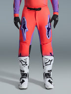 Alpinestars Pantaloni Supertech Scenz 2026, pantaloni da motocross, corallo acceso e viola, indossati con maglia coordinata, stivali bianchi e neri e guanti neri, con grandi loghi Astars viola sulle cosce e fibbia magnetica nera in vita, vestibilità atletica professionale per le competizioni
