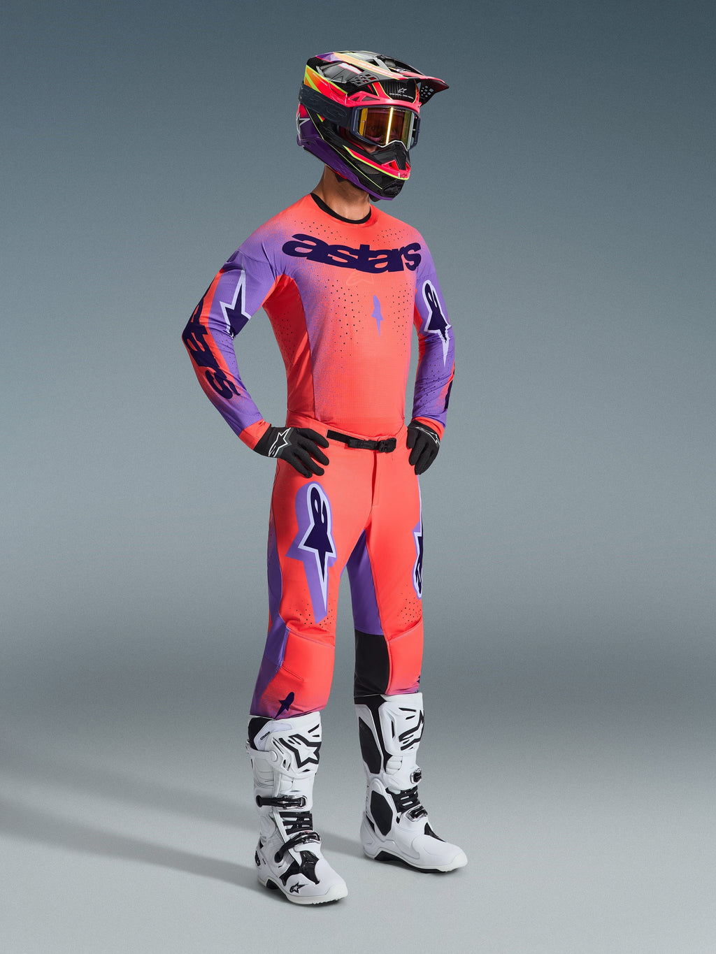 Pilota di motocross con abbigliamento da gara completo Alpinestars, pantaloni 2026 Supertech Scenz in Hot Coral Purple, maglia coordinata corallo e viola, casco nero e rosa, stivali bianchi e neri, kit professionale da gara off-road.