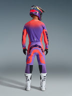 Alpinestars Pantaloni Supertech Scenz 2026, pantaloni da motocross, Hot Coral e viola, vista posteriore di un pilota di motocross che indossa un set completo di abbigliamento da gara, caratterizzati da un vivace design sfumato con loghi posteriori audaci, abbinati a maglia coordinata, casco e stivali bianchi.