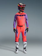 Pantaloni Alpinestars 2026 Supertech Scenz, pantaloni da motocross, corallo acceso e viola, indossati come set completo con maglia e casco coordinati, stivali bianchi e neri, con loghi viola e vestibilità atletica racing.
