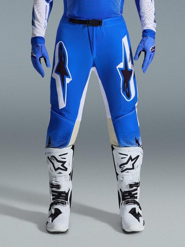 Alpinestars 2026 Supertech Scenz Pants, pantaloni da motocross, blu UCLA e bianco, indossati con guanti blu coordinati e stivali bianchi, con grandi grafiche bianche e nere sulle cosce, vestibilità atletica per prestazioni off-road