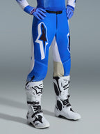 Alpinestars Pantaloni Supertech Scenz 2026, pantaloni da motocross, blu UCLA e bianco, indossati con stivali bianchi e neri e guanti blu, caratterizzati da grandi loghi grafici sulle cosce e pannelli bianchi, vestibilità atletica progettata per gare di motocross