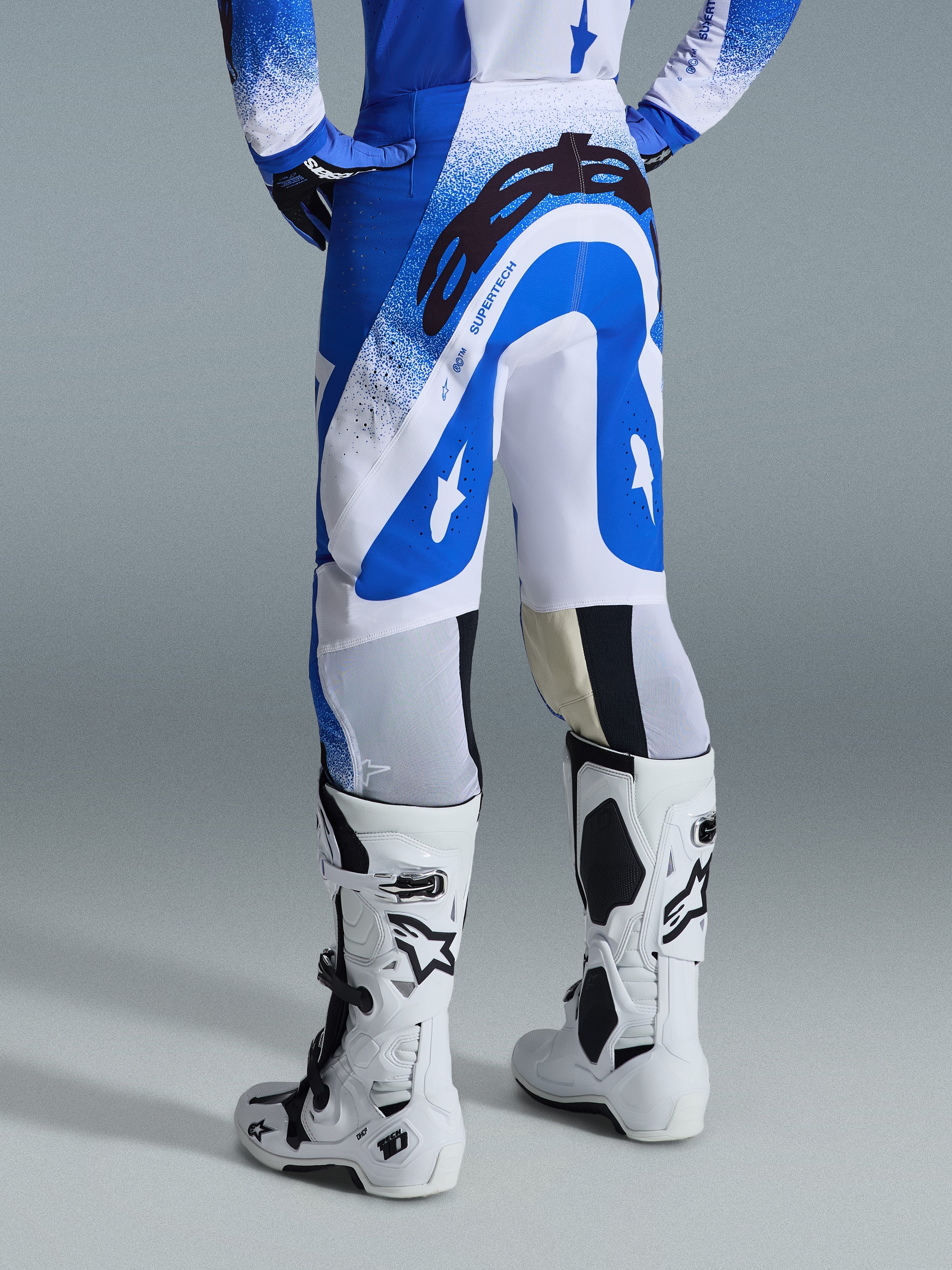 Alpinestars 2026 Supertech Scenz Pants, pantaloni da motocross, blu Ucla e bianco, vista posteriore con grande logo Astars nero sulla seduta e marchio 'SUPERTECH', indossati con stivali Tech 10 bianchi e maglia blu e bianca coordinata