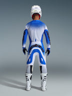 Pilota di motocross visto da dietro con abbigliamento completo da gara, pantaloni Alpinestars Supertech Scenz 2026 in blu Ucla e bianco, maglia coordinata blu e bianca con motivo puntinato, casco bianco, stivali bianchi e neri, outfit coordinato per competizioni professionali
