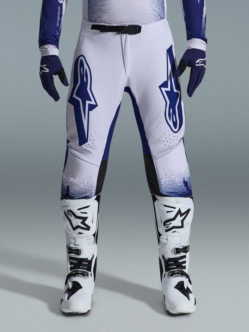Pantaloni Alpinestars Supertech Scenz 2026, pantaloni da motocross, grigio chiaro e blu scuro, indossati con stivali da motocross bianchi e neri e guanti blu scuro, caratterizzati da grandi loghi Alpinestars blu sulle cosce, fibbia magnetica SX1 e vestibilità atletica per corse fuoristrada