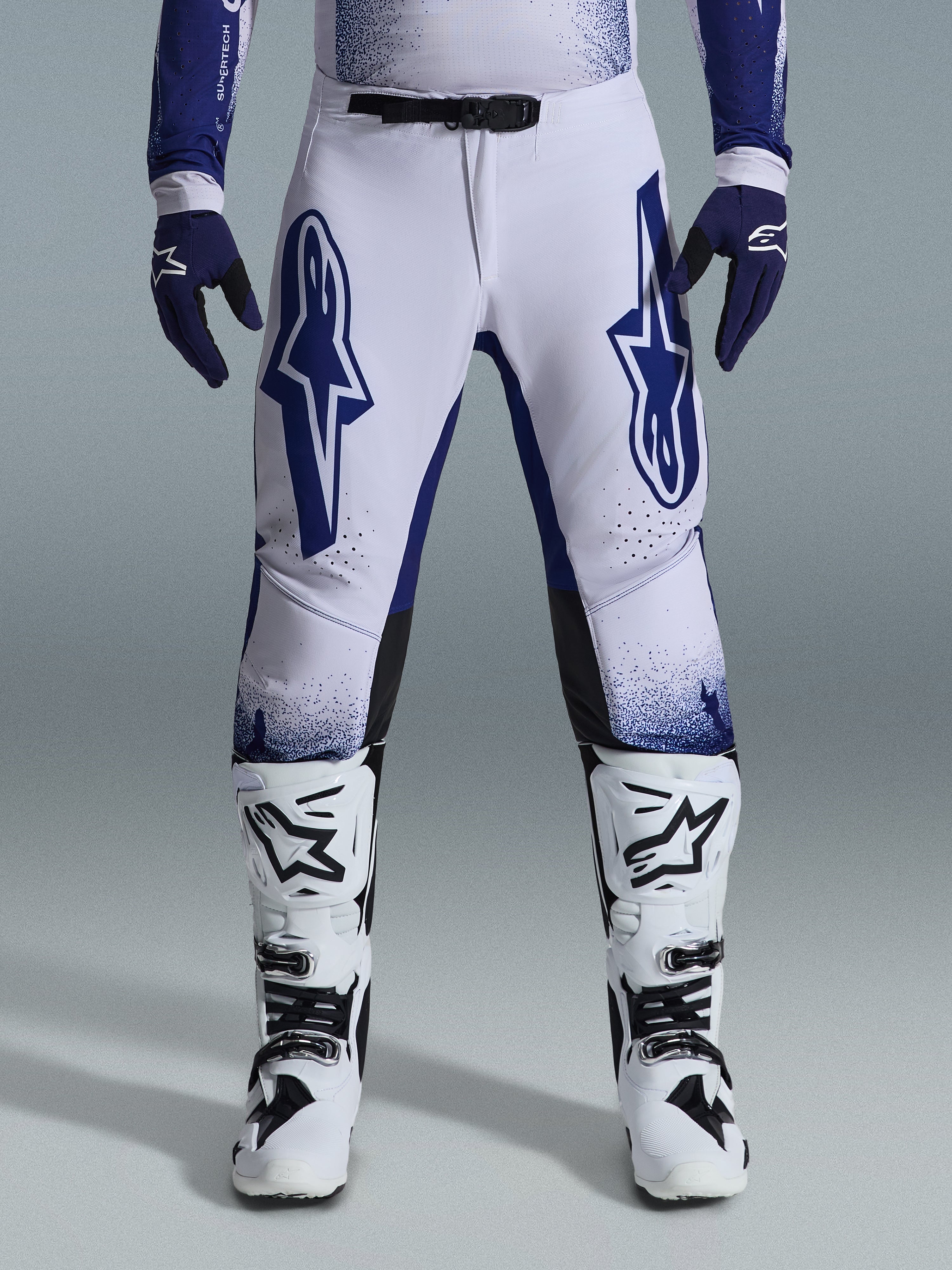 Pantaloni Alpinestars Supertech Scenz 2026, pantaloni da motocross, grigio chiaro e blu scuro, indossati con stivali da motocross bianchi e neri e guanti blu scuro, caratterizzati da grandi loghi Alpinestars blu sulle cosce, fibbia magnetica SX1 e vestibilità atletica per corse fuoristrada
