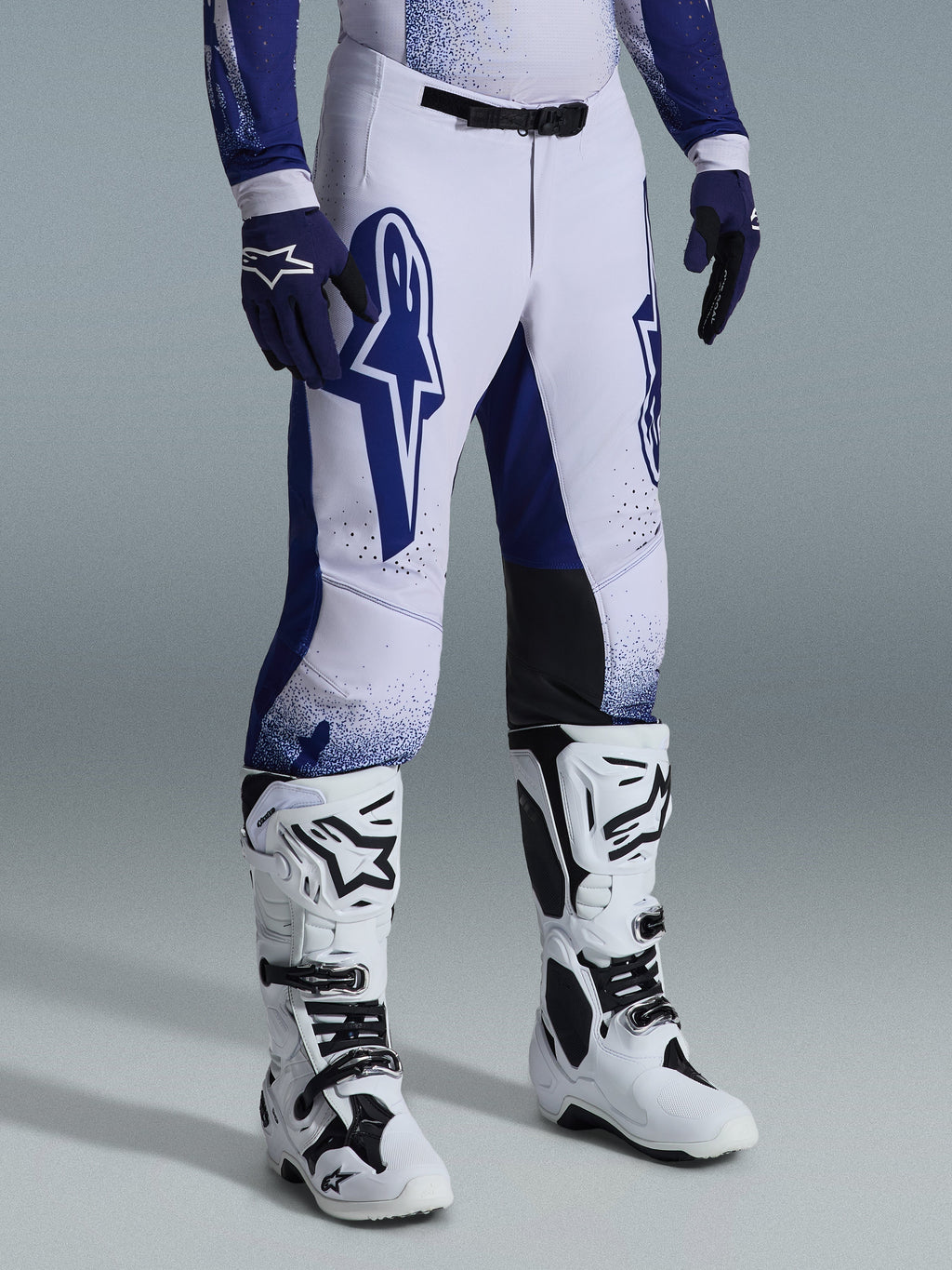 Alpinestars Pantaloni Supertech Scenz 2026, pantaloni da motocross, grigio chiaro e blu scuro, indossati con stivali da motocross bianchi e guanti blu, caratterizzati da grandi loghi Alpinestars blu sulle cosce, pannelli neri sulle ginocchia e design grafico puntinato, abbigliamento professionale da gara fuoristrada