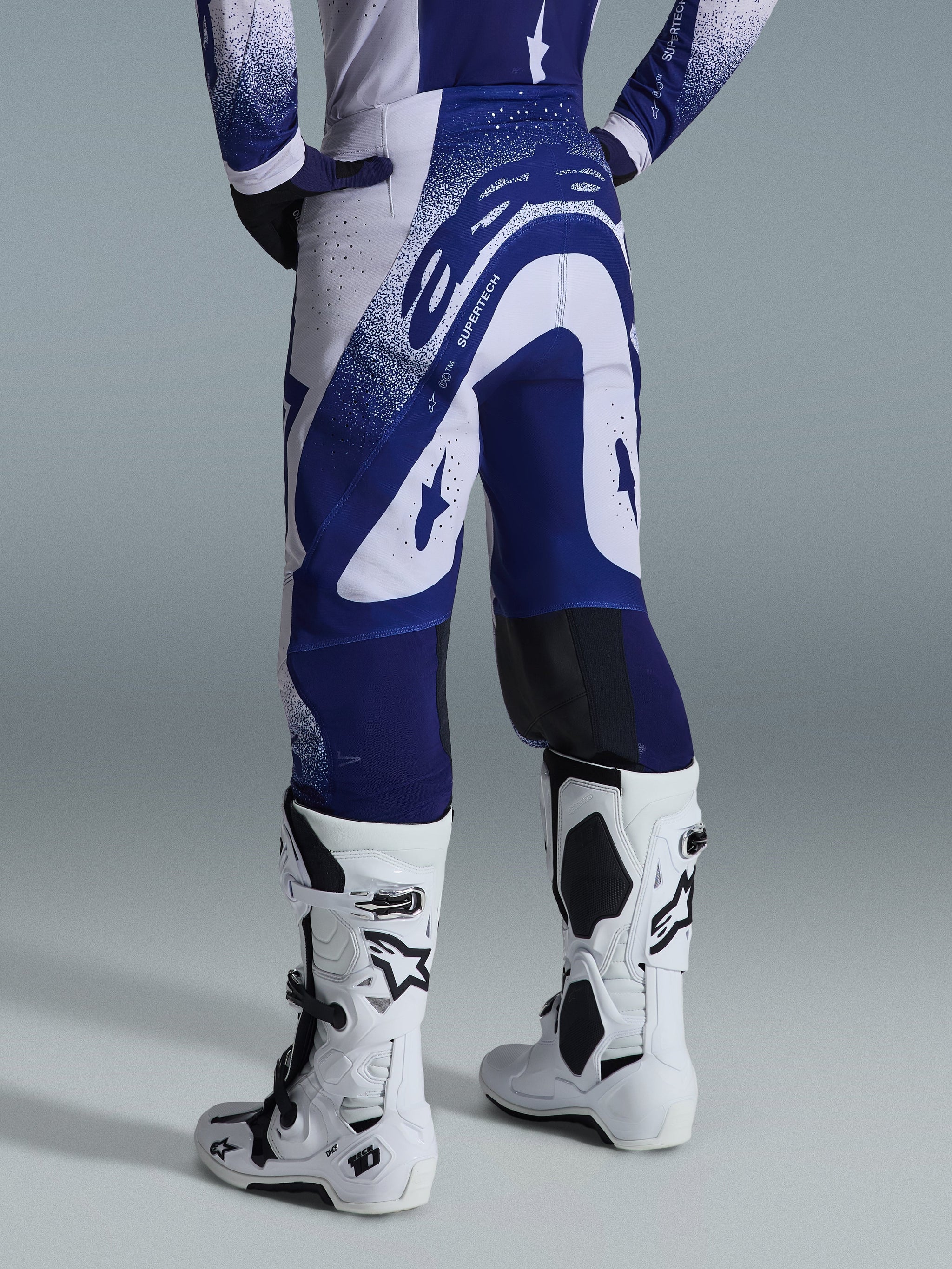 Alpinestars Pantaloni Supertech Scenz 2026, pantaloni da motocross, grigio chiaro e blu scuro, vista posteriore indossati con maglia coordinata e stivali bianchi, con grafiche blu vivaci e accenti punteggiati, vestibilità atletica per gare fuoristrada