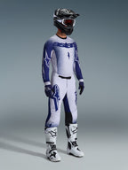 Pilota di motocross con pantaloni Supertech Scenz 2026 in grigio chiaro e blu scuro, maglia blu e bianca coordinata con logo 'astars' ed effetto spray, casco nero con occhiali bianchi, stivali da motocross bianchi e neri, completo professionale da gara fuoristrada.